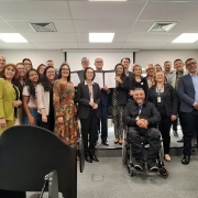 Fotografia de grupo feita em uma sala ampla e iluminada, semelhante a uma sala de reuni&otilde;es ou audit&oacute;rio. Cerca de duas dezenas de pessoas est&atilde;o reunidas no centro da imagem, posicionadas em duas fileiras e voltadas para a c&acirc;mera. Algumas pessoas, no centro do grupo, seguram folhas de papel brancas abertas, representando os termos assinados pelas pessoas presentes e voltadas para a fotografia.

Na parte central da frente, uma pessoa est&aacute; em uma cadeira de rodas, com as m&atilde;os unidas &agrave; frente do corpo, que &eacute; o presidente da FADERS, Marquinhos Lang. Ao redor, as demais pessoas est&atilde;o em p&eacute;. O grupo usa roupas variadas, incluindo ternos e blazers em tons escuros, al&eacute;m de pe&ccedil;as nas cores verde, marrom, branco e preto. Algumas pessoas utilizam crach&aacute;s.

Em primeiro plano, v&aacute;rias poltronas pretas vazias ocupam a parte inferior da fotografia e encobrem parcialmente a vis&atilde;o do grupo. &Agrave; direita, uma pessoa est&aacute; sentada em uma dessas poltronas, de costas e de perfil para a c&acirc;mera, segurando um objeto semelhante a um celular.

A sala possui carpete cinza, paredes claras e teto com lumin&aacute;rias retangulares e sa&iacute;das de ventila&ccedil;&atilde;o. Nas laterais superiores existem caixas de som pretas. &Agrave; esquerda h&aacute; grandes janelas, por onde entra luz natural. Ao fundo, atr&aacute;s do grupo, encontra-se uma grande tela de proje&ccedil;&atilde;o branca recolhida. &Agrave; direita, um monitor grande fixado &agrave; parede exibe uma interface de computador com textos pequenos, que n&atilde;o podem ser lidos na fotografia.

A composi&ccedil;&atilde;o concentra a aten&ccedil;&atilde;o no grupo reunido e, especialmente, nos documentos exibidos no centro.